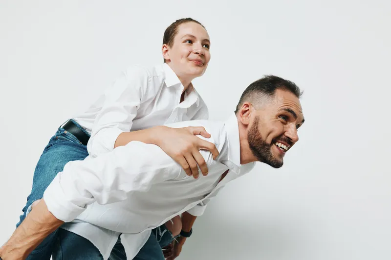 Two professionals, a confident man and his teammate, share a playful piggyback moment on a clean white background, showcasing teamwork, trust, and positive energy in a corporate setting.