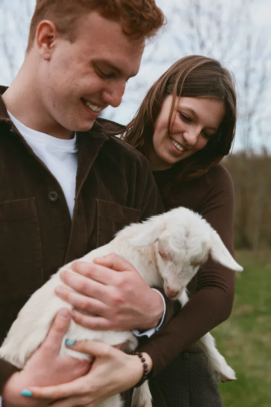 A casual couple gently holds a young goat kid in an outdoor field, smiles warm, sunlight bright. A lifestyle moment showcasing care, companionship, and rural charm in everyday animal bonding.