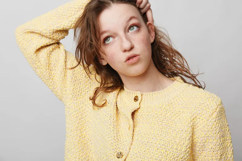 Young woman in yellow knitted sweater looks up thoughtfully with hand on head. Studio portrait on grey background. Concept of contemplation, doubt, and casual fashion.