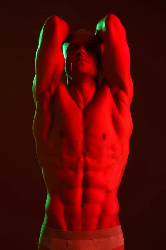 Athletic man posing dramatically under vibrant red and green lighting, showcasing a strong physique. This image captures power and intensity in a unique style.
