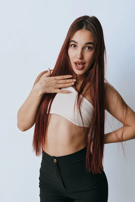 Young woman with long hair posing with surprised expression and hand on chest wearing crop top and black pants isolated on white background in studio.