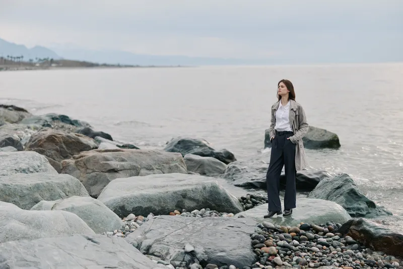 Young woman stands on rocky shore near calm sea, wearing casual outfit with jacket and trousers. Overcast sky creates peaceful and contemplative mood by the water. Concept of solitude and nature.