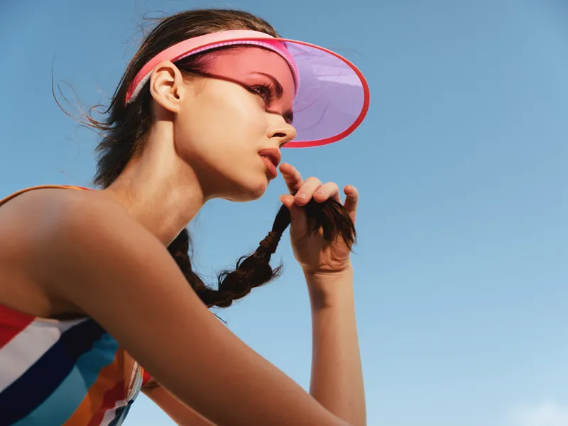 Close-up portrait of a female athlete wearing a pink visor, braid in motion, and bold sunlit sky behind. The image emphasizes concentration, athleticism, and modern sportswear.