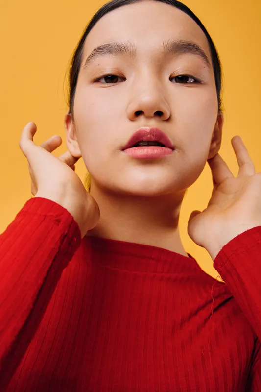 Woman with smooth skin and natural makeup posing with hands near face against yellow background. Young female portrait in red sweater with confident expression and soft lighting.