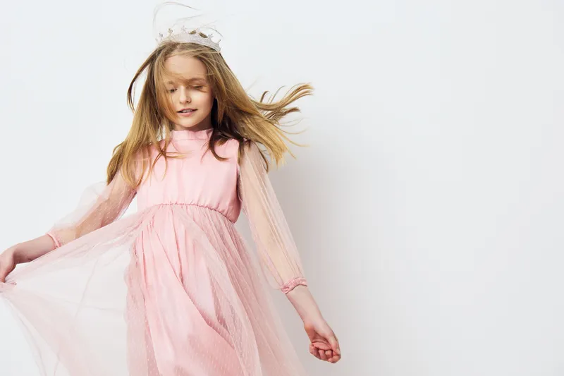 Young girl in a light pink dress with flowing tulle sleeves, displaying joy and innocence against a bright white background, perfect for fashion or childhood concepts