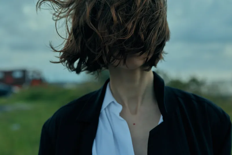 Wind swept hair and exposed neck in an outdoor setting with a blurred countryside field, a casual jacket over an open white shirt, creating a moody, enigmatic fashion moment that hints at mystery