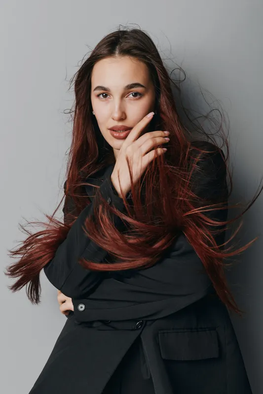 Young woman with long red hair wearing black jacket posing against gray background with hand near face looking confidently at camera in studio portrait with natural expression.