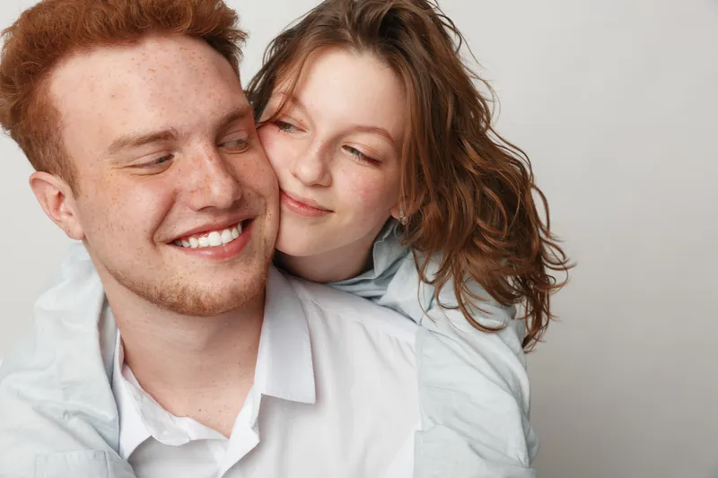 Happy young couple with red hair and casual clothes smiling and embracing each other against a plain background. Concept of love, affection, friendship, and positive emotions.