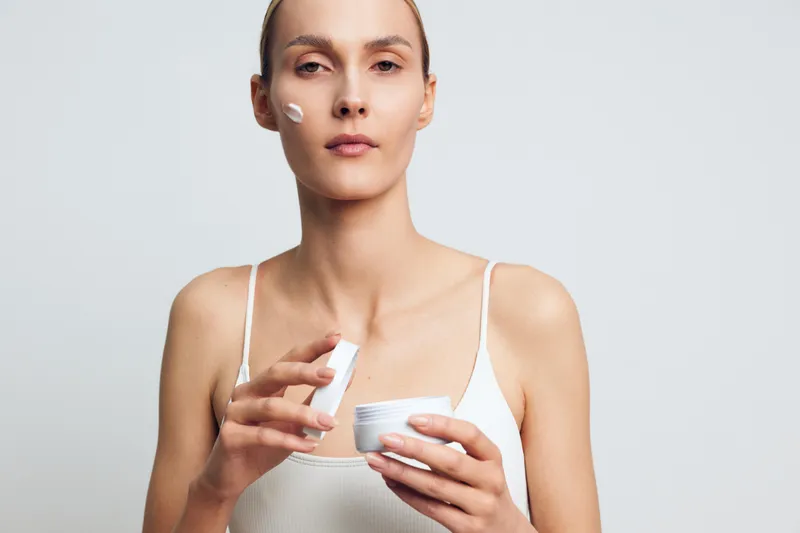 Young woman applies face cream with fingers holds jar of moisturizer skincare routine natural beauty isolated on white background minimalistic style