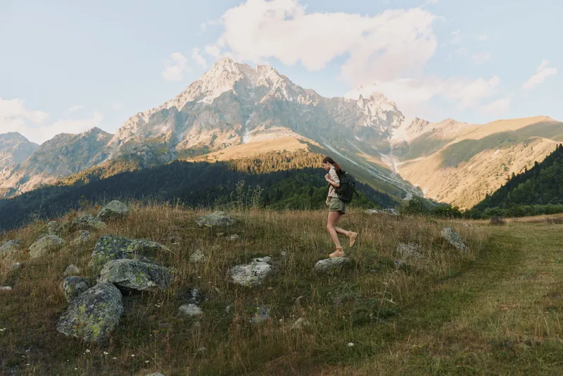 Young woman with backpack hikes through a grassy mountain valley under a partly cloudy sky, enjoying the peaceful natural landscape and fresh air.