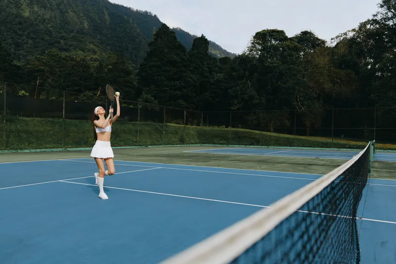 Young woman playing tennis outdoors, wearing sporty attire, with a joyful expression, on a blue court surrounded by greenery, ideal for sports and fitness concepts