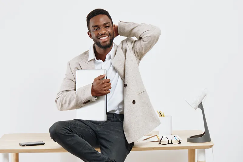 Confident smiling man in casual beige jacket sitting at desk holding a tablet device in a bright minimalist office background with workspace items. People portrait concept.