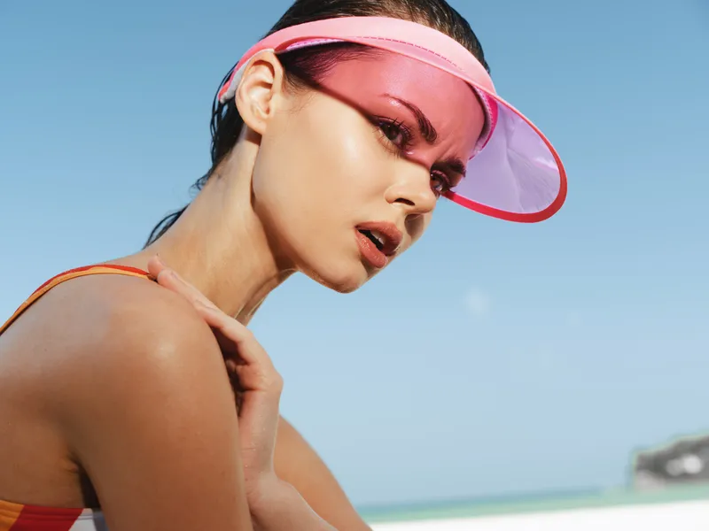 Close up of a sunlit woman wearing a pink visor, glowing skin and bold makeup, gazing toward the sea with a thoughtful, captivating expression.