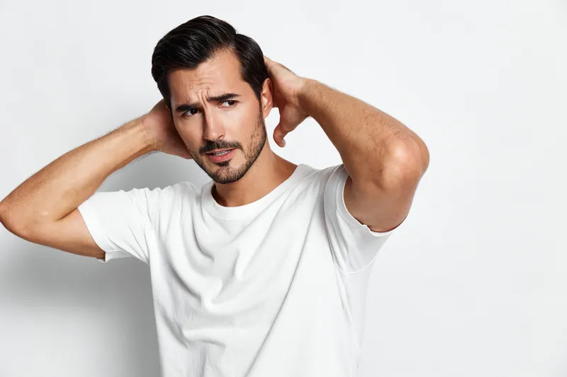 Handsome young man casual wearing white t-shirt looking at camera with thoughtful expression, holding hands behind head, standing against light grey background.