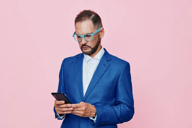 A focused business professional man in a blue suit stands against a solid pink background, projecting confidence and concentration as he checks his device.