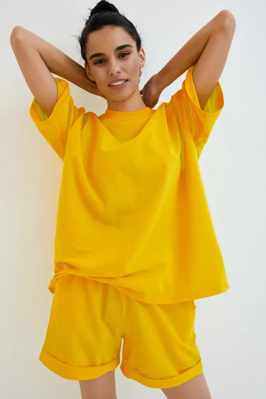 young woman in yellow casual outfit posing with hands behind head against plain wall, relaxed mood, natural light, summer clothing style