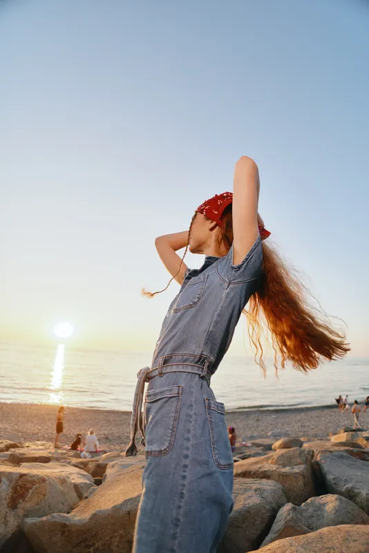 lifestyle woman in modern boho-western denim street style relaxing at sunset by the beach with warm film color tones and natural sunlight