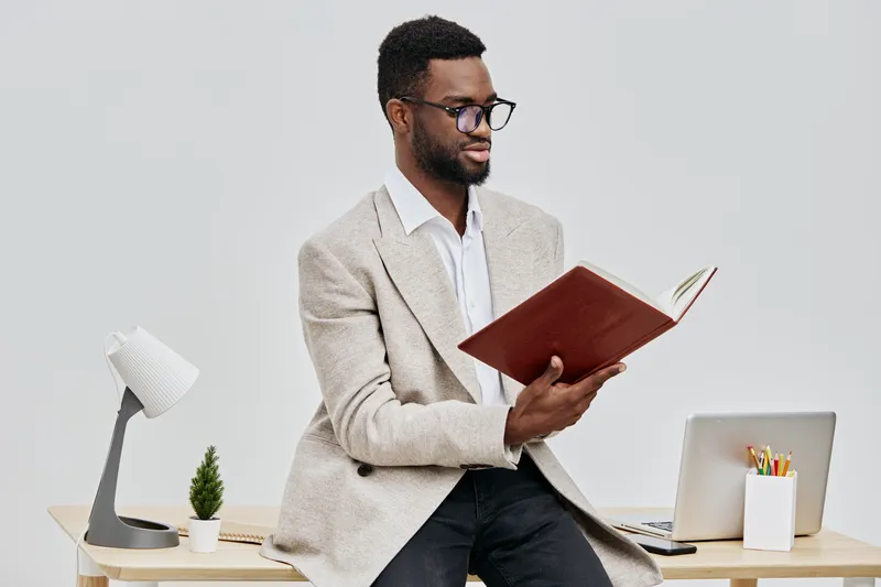 Thoughtful man in beige cardigan sitting on desk and reading a book, workspace with laptop and stationery on a plain white background, studio shot. Business and education concept