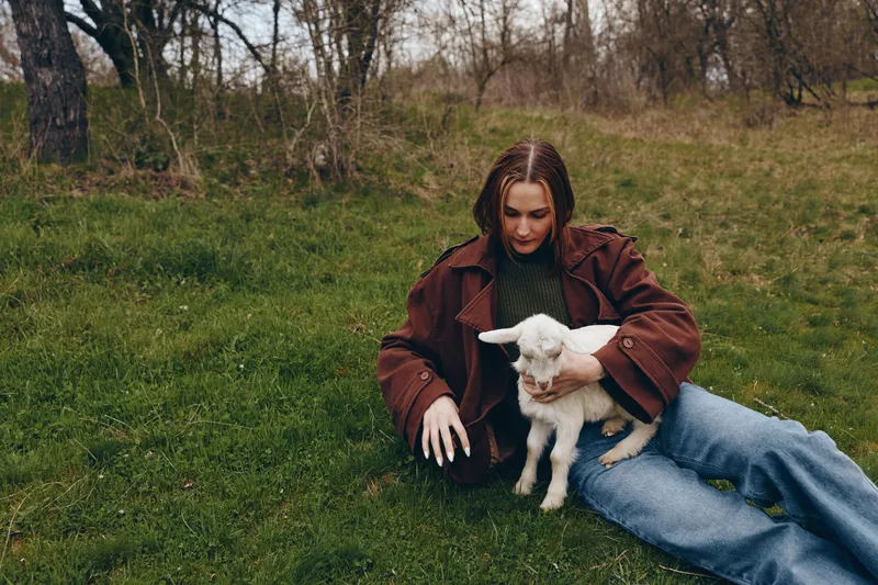 A woman sits on green grass in a park, gently cradling a small white goat kid, enjoying a tranquil outdoor moment, warm companionship, and a simple lifestyle scene.