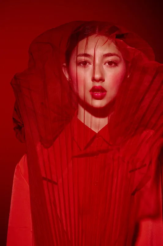 Creative portrait of a young fashion model in bright red dress and dramatic sheer veil. Studio lighting highlights her intense expression. Concept for beauty, fashion, art.