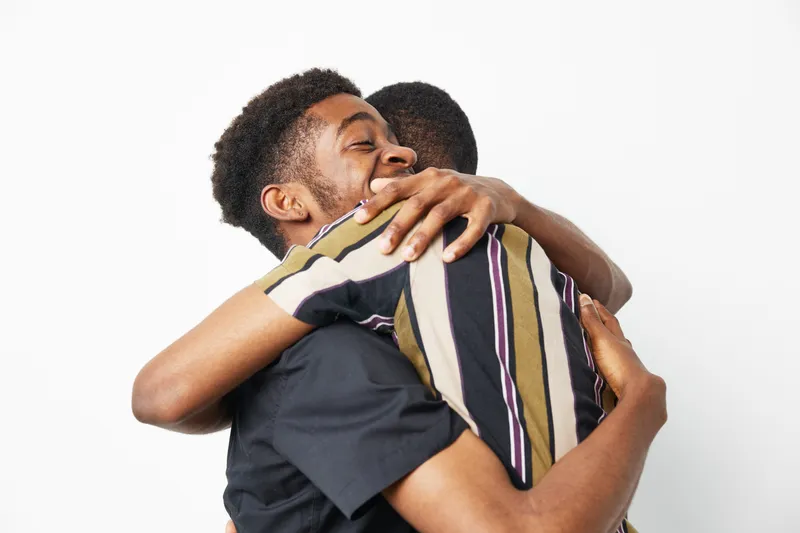 Joyful black men sharing a warm embrace, expressing friendship, trust, and comfort, captured in a bright setting with a clean background.