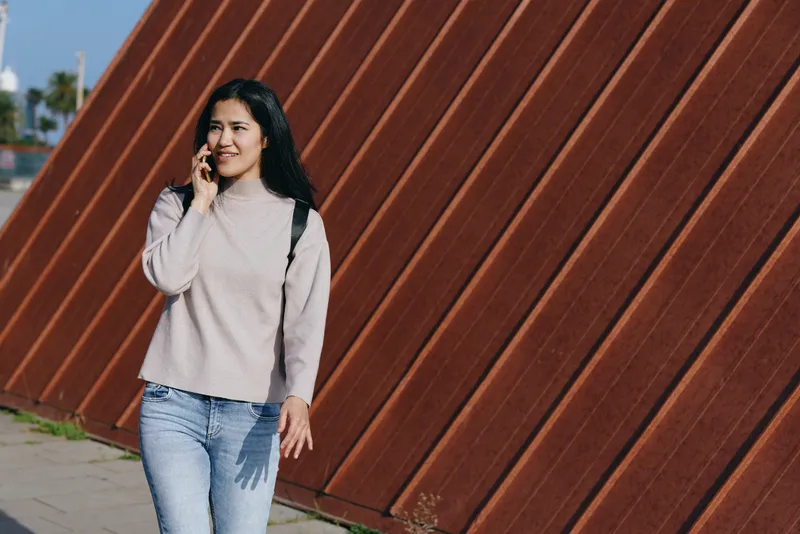 Casual woman in sweater and jeans talks on a smartphone while walking beside a rust diagonal wall in an urban outdoors setting, capturing everyday communication and contemporary street fashion