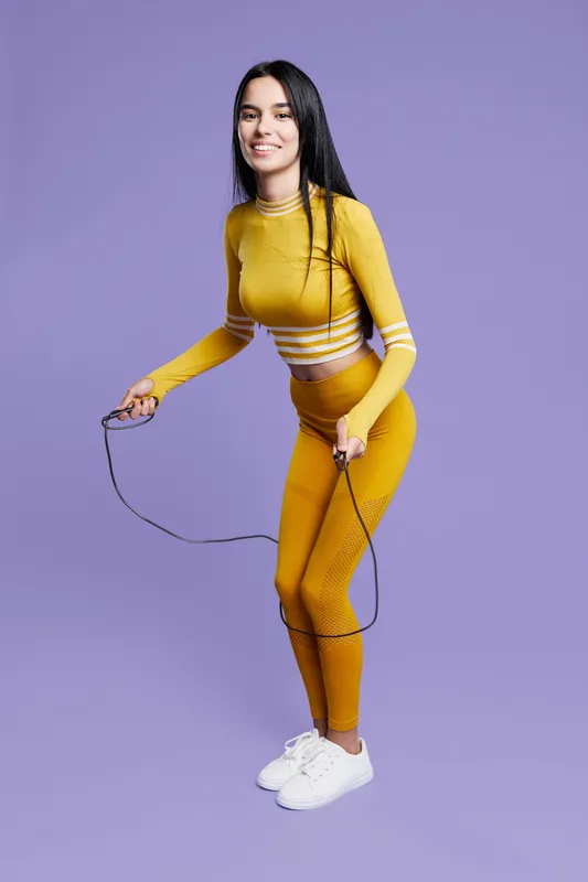 Woman sport fitness wearing bright yellow activewear jumps rope with smile, showing energy, health, and workout motivation against purple background in studio setting.