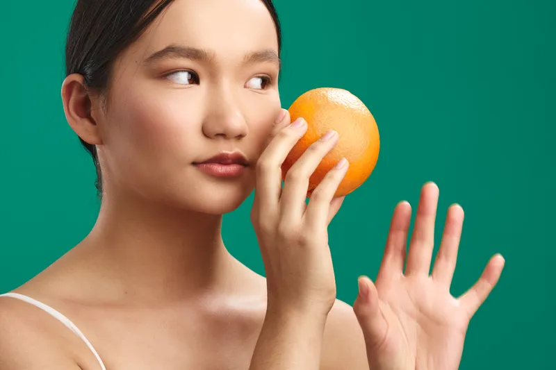 asian woman holds an orange near her face against a vivid green backdrop, calm expression, soft light, playful yet refreshing concept emphasizing health and vitality.