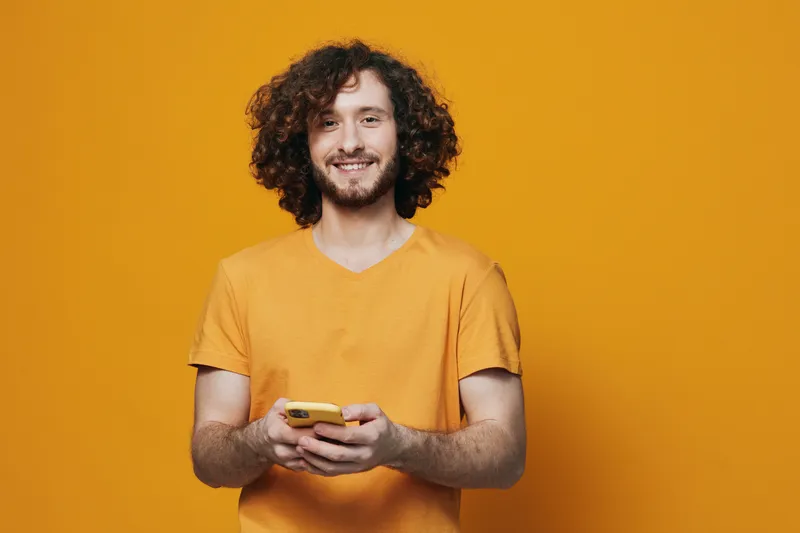 Smiling young man using smartphone, wearing a vibrant orange t shirt against a bright orange background, embodying a joyful and modern lifestyle