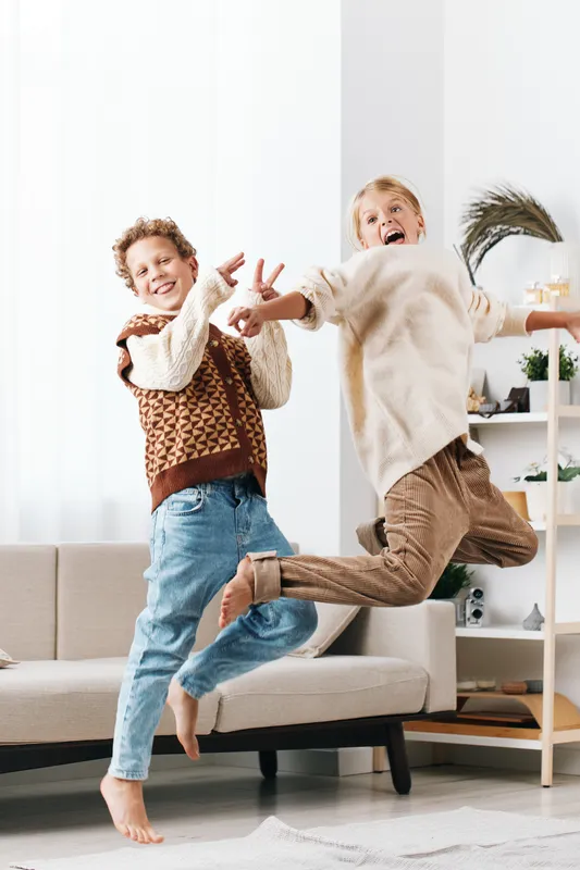 Authentic playful scene of school aged children at home, capturing a genuine lifestyle moment as they jump and laugh, reflecting everyday joy and friendship.