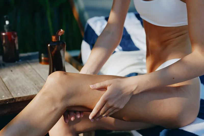 A woman in white underwear applies lotion to her leg in a sunlit room with wooden furniture. The image represents self-care, skincare, and wellness in a relaxed lifestyle setting.