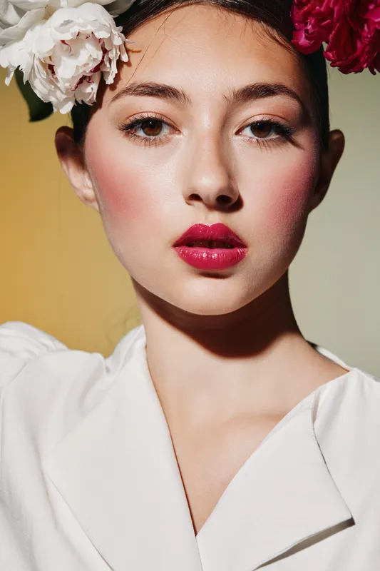 Young woman with creative bright makeup and red lips wears a floral headpiece and white outfit. Close-up portrait on muted yellow background. Concept of beauty fashion editorial.