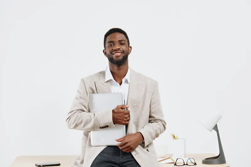 Confident smiling man in business casual attire holding a silver laptop while standing in modern office setting with minimal decor and white background. Professional, success, executive, tech, workpla