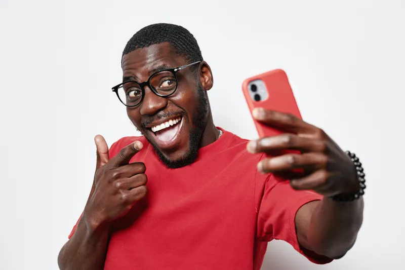 African American man with dark skin wearing glasses and red shirt taking selfie with smartphone smiling and pointing finger on white background in studio portrait.