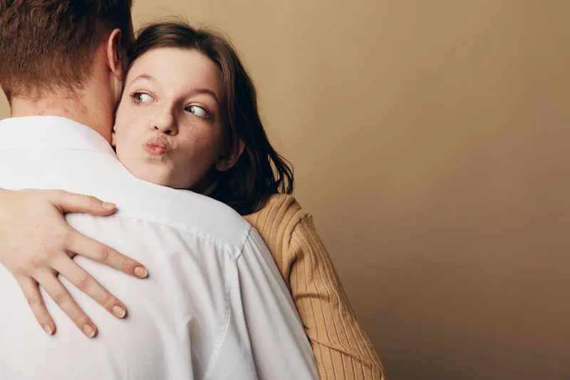 Young woman in beige sweater hugs man in white shirt, making playful face. Intimate moment with warm brown background. Concept of affection, relationship, and casual lifestyle.