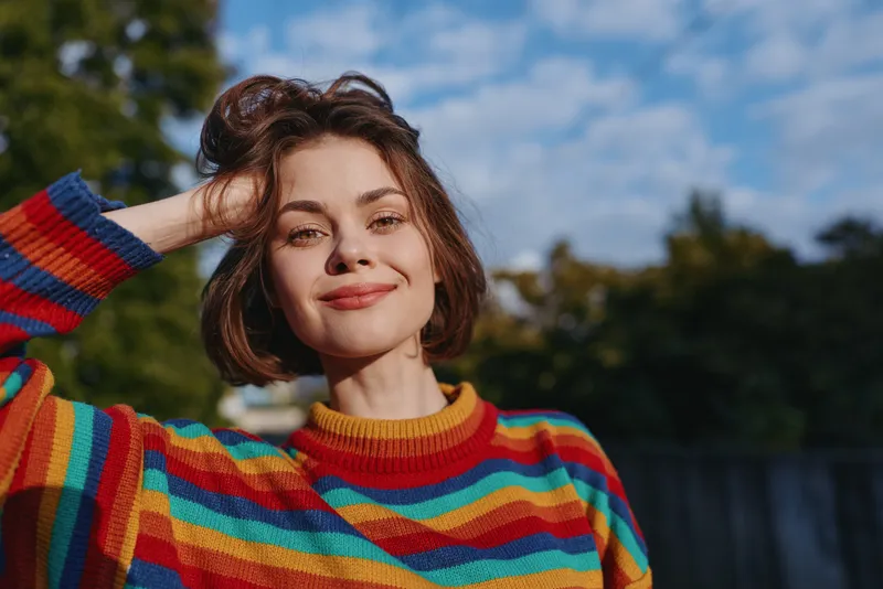 Woman portrait smile in colorful sweater outdoors with sunlight on face, short hair and relaxed pose, natural expression and shallow depth of field for lifestyle and fashion.