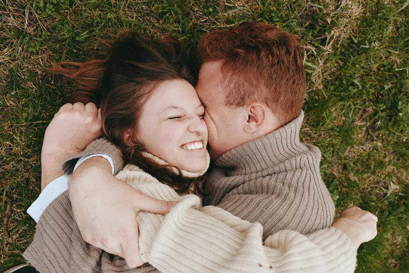 Smiling couple lying on grass, embracing in cozy sweaters. Outdoor scene captures warmth, affection, and intimate connection under natural light on a soft lawn with a gentle breeze and sunlight.