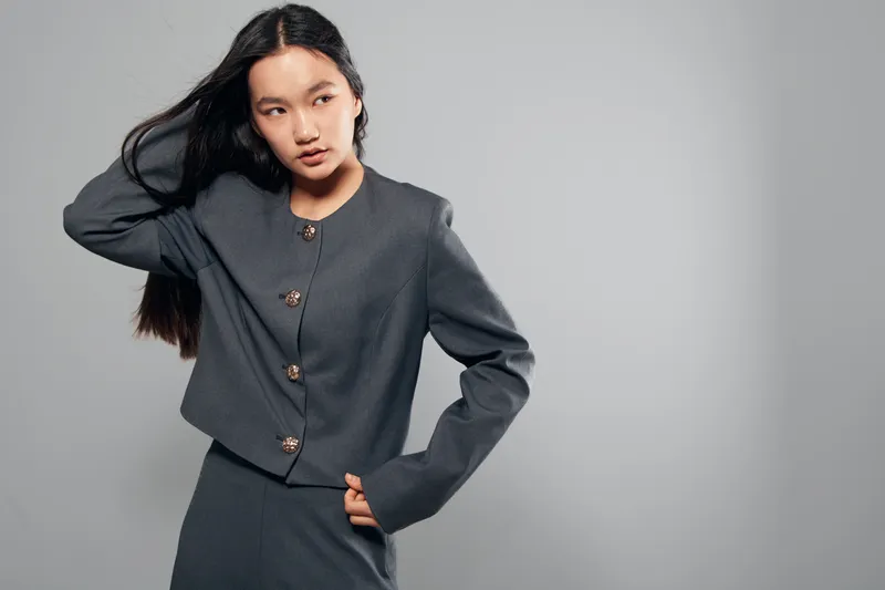 Woman with long black hair wearing gray stylish jacket and skirt posing with hand on hip against neutral background. Fashion model showing contemporary outfit and confident posture.