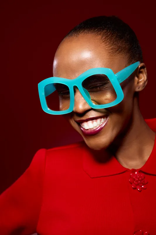 Smiling black woman with dark skin wearing large blue glasses and a red top. Portrait with closed eyes on red background. Concept of fashion, joy, and vibrant style.