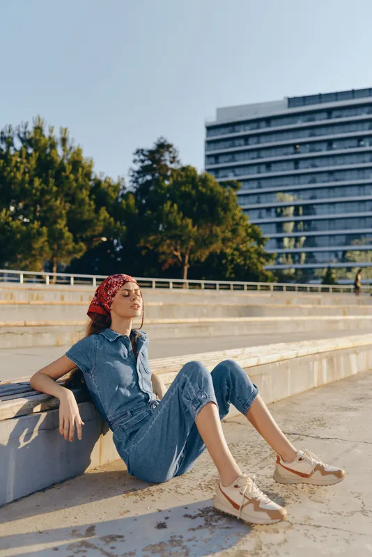 Lifestyle woman in modern boho-western denim street style, sitting relaxed outdoors with sunglasses and a red bandana, captured in warm film color tones.
