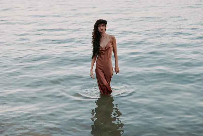 A solitary woman stands waist deep in calm water wearing a flowing rust dress, soft light highlights her silhouette, reflective surface adds serenity and depth.