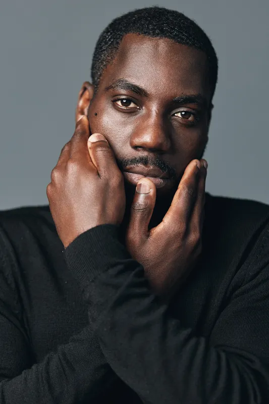 African man with dark skin in creative conceptual visual style posing with hands on face against gray background. Portrait of thoughtful male model wearing black sweater in studio lighting.