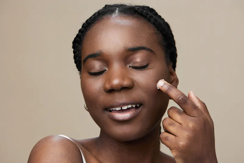 Young Black woman applying skincare product, smiling with closed eyes, enjoying beauty routine against neutral background