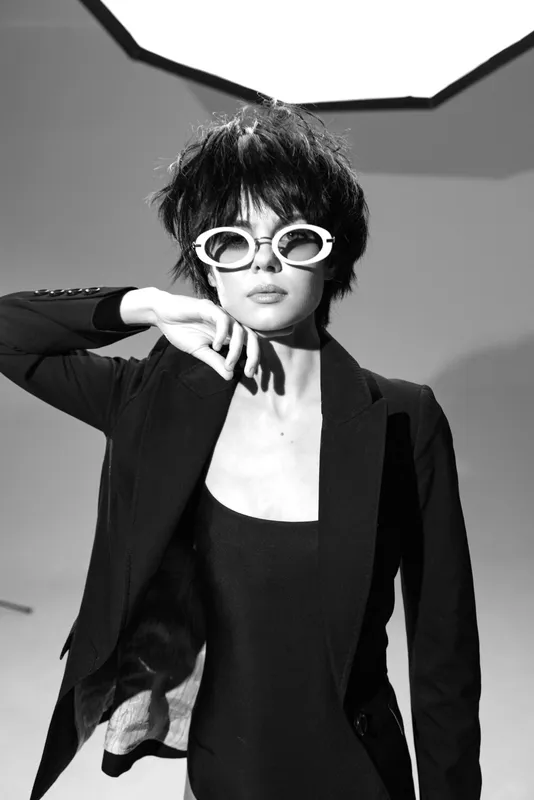 Black and white portrait of a stylish woman wearing sunglasses and a jacket in a studio setting.