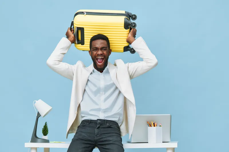 Happy young African American man in white shirt and blazer smiling and holding a yellow suitcase above his head, standing in a bright modern office environment with a desktop, lamp, plants, and statio