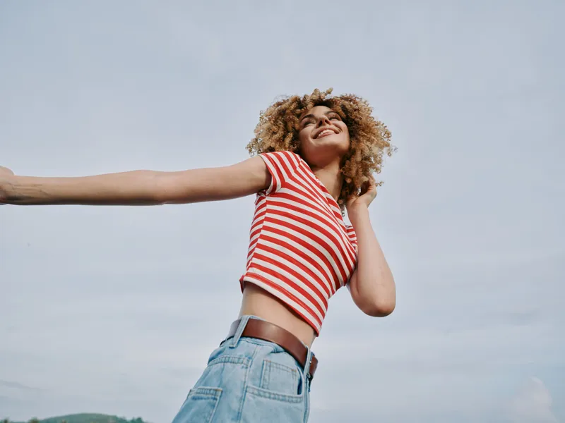Joyful figure stretches arms wide under a pale sky, wearing a red striped crop top and denim shorts, capturing carefree summer vitality and open, uplifting energy.