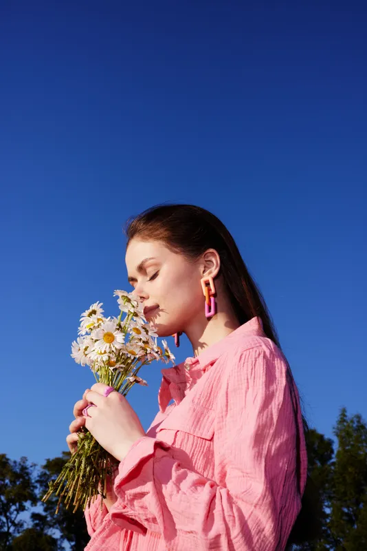 Young woman holding daisies against a clear blue sky, wearing pink clothing and colorful earrings, radiating joy and warmth in a vibrant outdoor setting