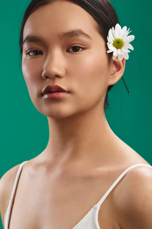 Asian woman with a white flower tucked behind her ear gazes softly at the camera against a green backdrop, presenting a serene portrait that emphasizes natural beauty and calm elegance.