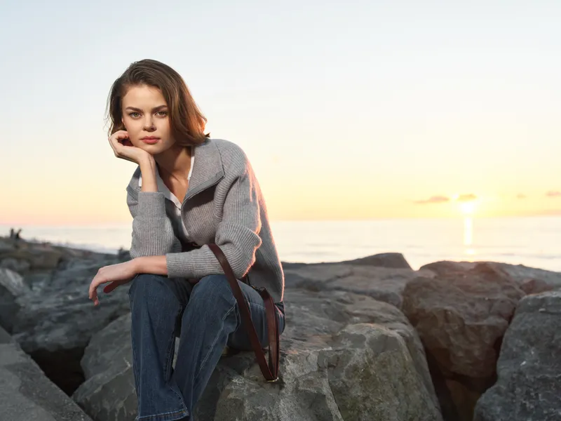 A thoughtful woman sits on rocky shore at sunset, resting her chin on her hand. She wears a gray sweater and jeans, exuding calm and quiet reflection.