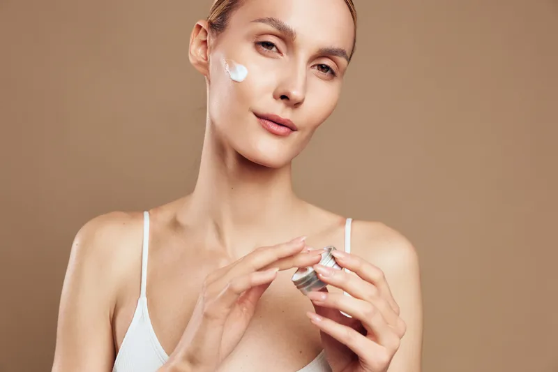 Woman skincare cream face beauty treatment spa hygiene, moisturize wellness daily care, young portrait, white cream on cheek, gentle smile, beige studio light soft fresh glow